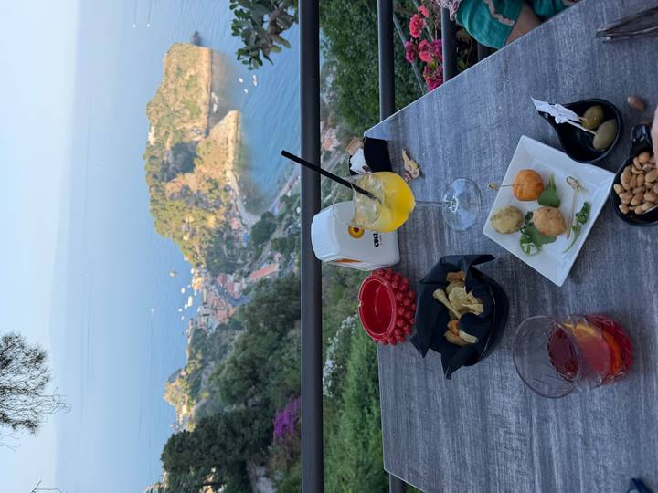 Drinks and Sicilian snacks on a terrace table overlooking a picturesque bay dotted with boats far below.