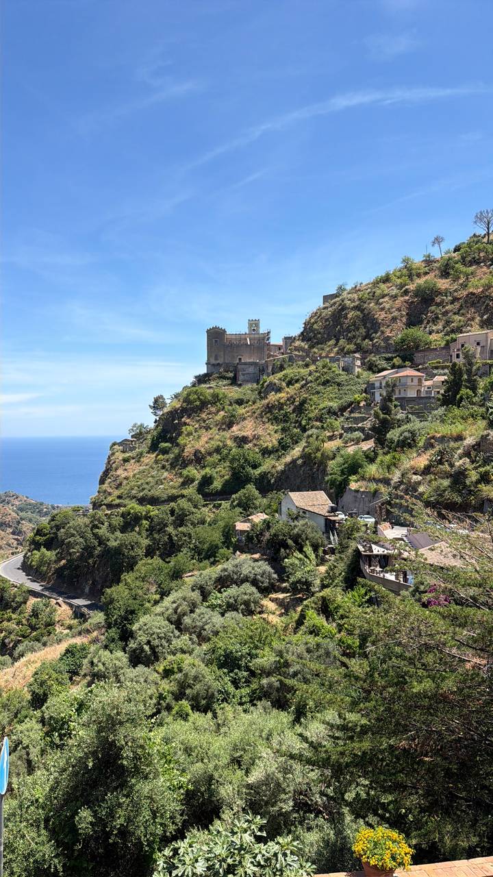 Historic hilltop castle and clustered houses perch above the Mediterranean coast with deep blue sea below.