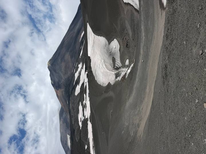 Volcanic slopes of Mount Etna with dark ash fields and lingering snow patches beneath dramatic clouds.