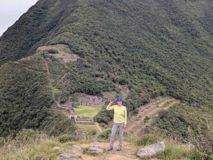 A trekker poses with a peace sign overlooking the expansive Inca ruins of Choquequirao set in lush mountains.