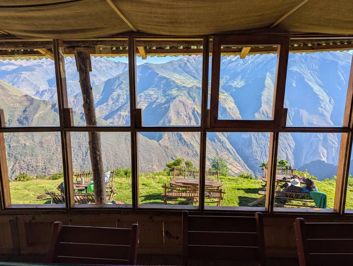Panoramic mountain scenery framed by a wooden lodge window showing grassy terrace tables high above an Andean valley.