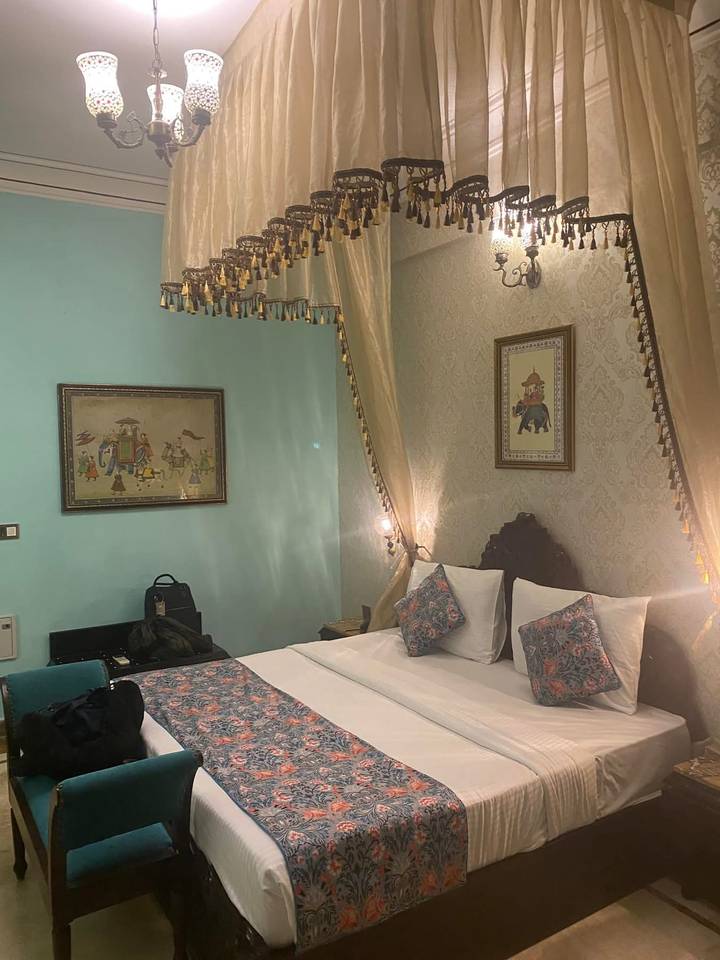 A richly decorated hotel bedroom with pastel walls, framed artwork, a canopy, and patterned pillows on a double bed.