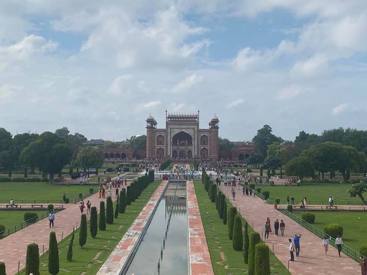 A broad view of the grand gateway to the Taj Mahal complex with long reflecting pools and manicured gardens bustling with visitors.