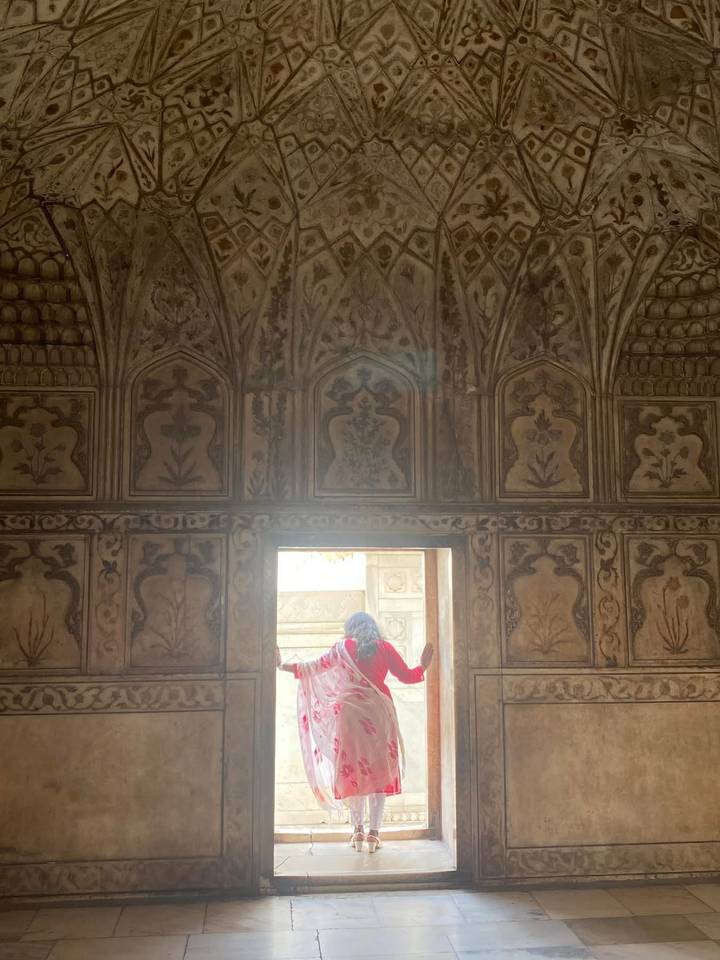 A woman in a bright red outfit stands in an ornate stone chamber, looking out an arched doorway.