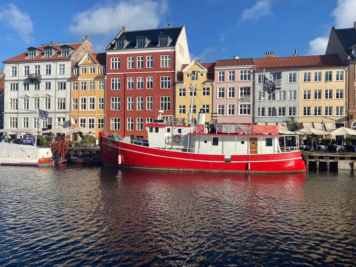 A vivid red boat floats along Nyhavn with rows of pastel canal houses under clear blue skies.