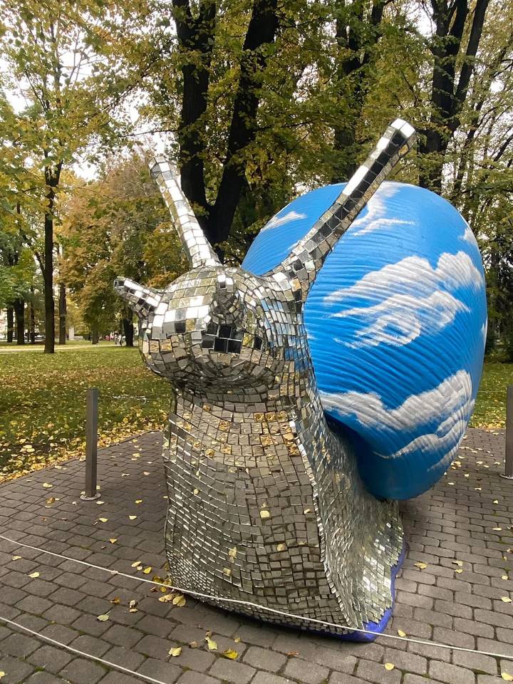 A large mirrored snail sculpture with sky-cloud patterns stands in a leafy city park.