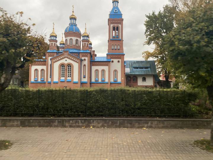 Ornate blue-domed Orthodox church with gold spires seen from across a hedge on a cloudy day.