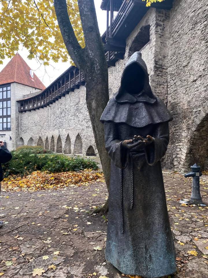 Bronze hooded monk statue in front of stone medieval wall and autumn leaves.