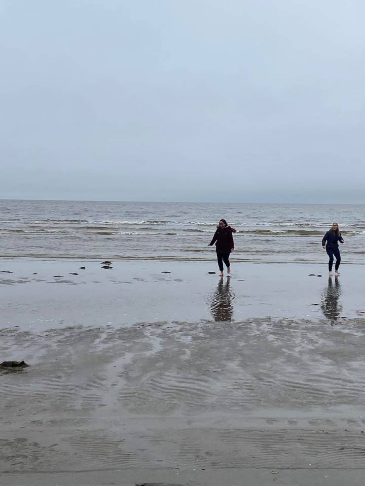 Two young women walk barefoot in the shallow water on a grey deserted beach.