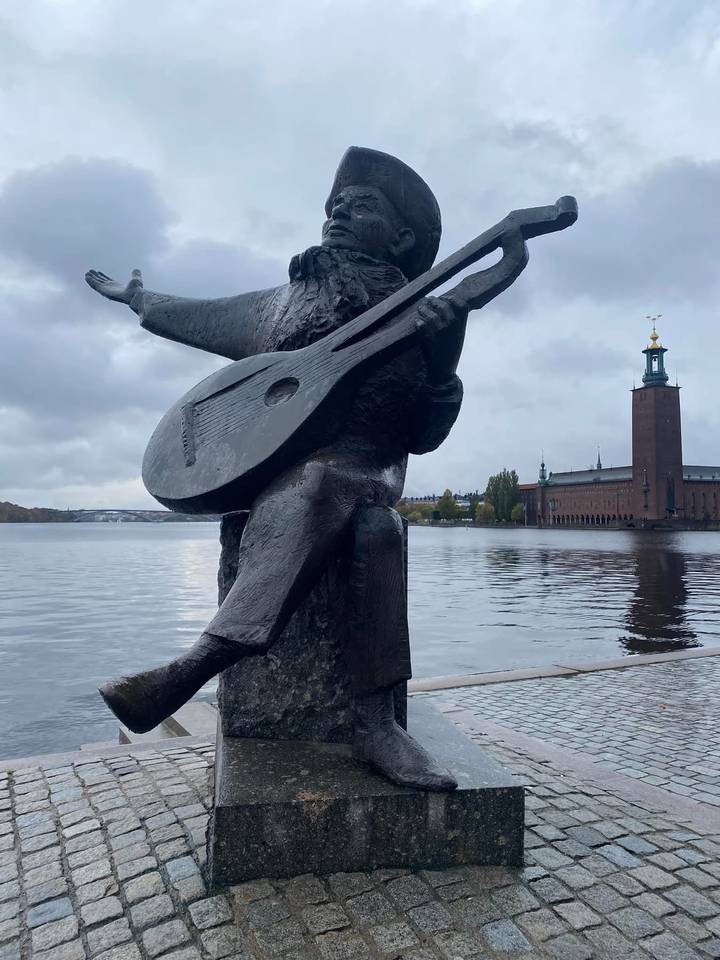 Statue of a guitarist overlooking the water with Stockholm City Hall in the background under cloudy skies.