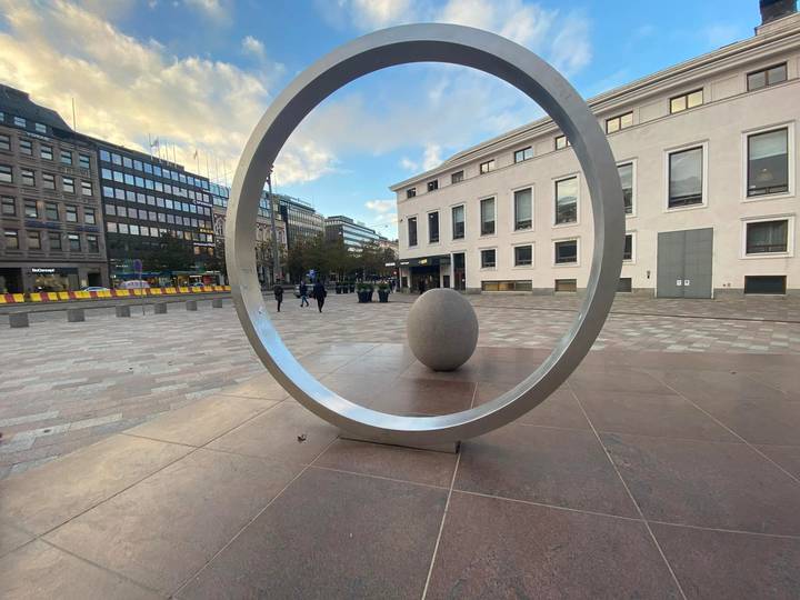 Modern outdoor sculpture of a metal ring framing an egg-shaped stone in a city plaza.