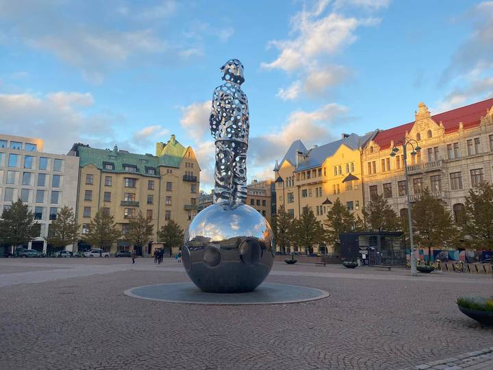 Reflective sphere sculpture topped by a perforated human figure in a European square at golden hour.