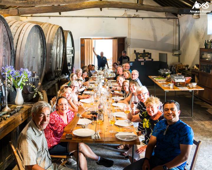 Large tour group enjoying a wine tasting meal around a long rustic table in a cellar.