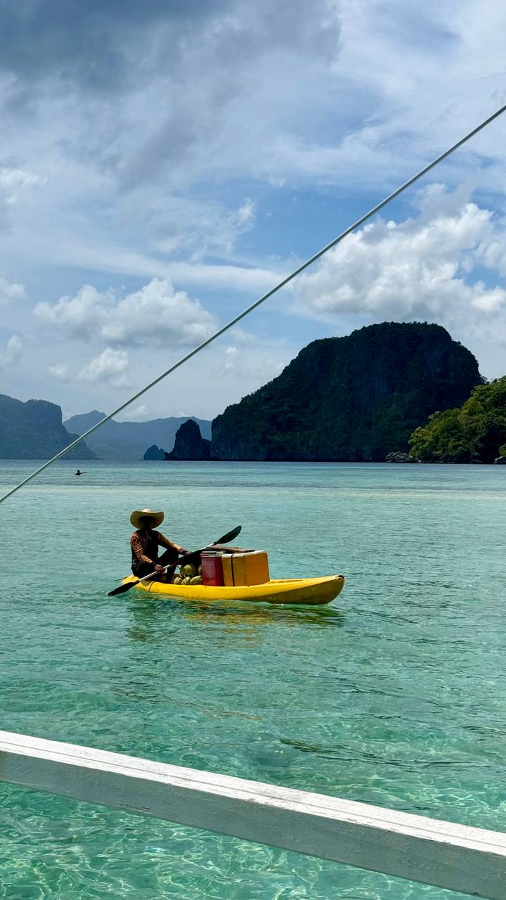 Man in straw hat paddling a yellow kayak laden with goods across serene jade waters with karst cliffs behind.