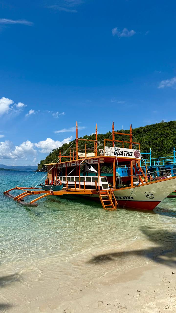 Bright wooden outrigger tour boat moored in clear blue water with green forested hills and blue sky.