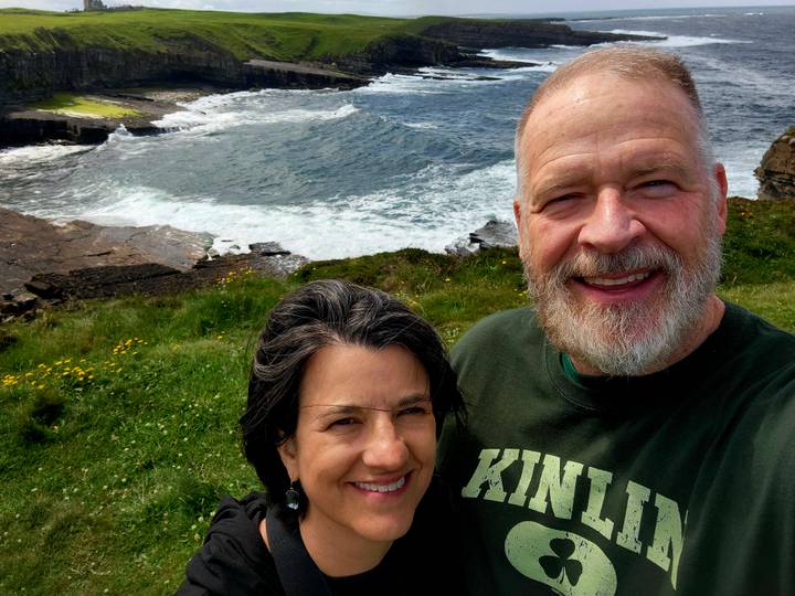 Couple smiling with rugged coastline and crashing waves behind them