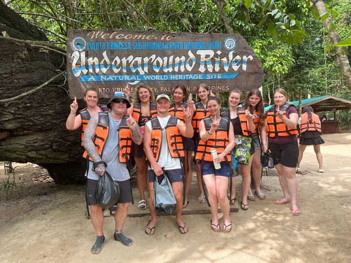 Group of smiling travellers wearing orange life vests posing in front of the Puerto Princesa Underground River entrance sign.