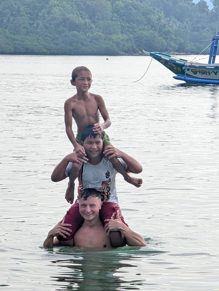 Three playful boys enjoying the water, one sitting on another’s shoulders against a calm sea backdrop.