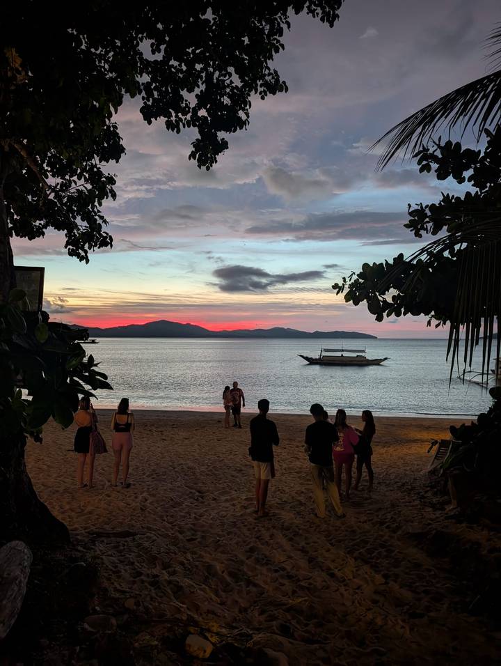 Silhouettes of beachgoers watching a vivid pink and orange sunset over calm water with an outrigger boat anchored offshore.