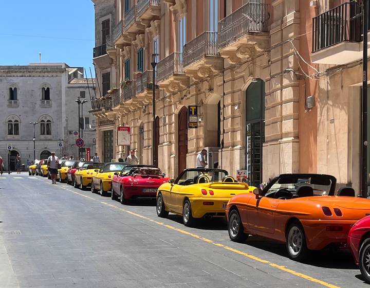 Row of colorful convertible sports cars parked along a historic Sicilian street.