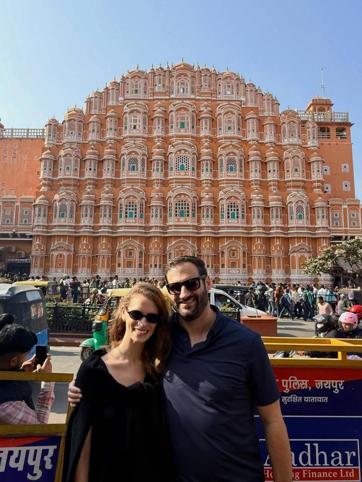 Couple standing before the ornate pink façade of Jaipur’s Hawa Mahal amid bustling street life.