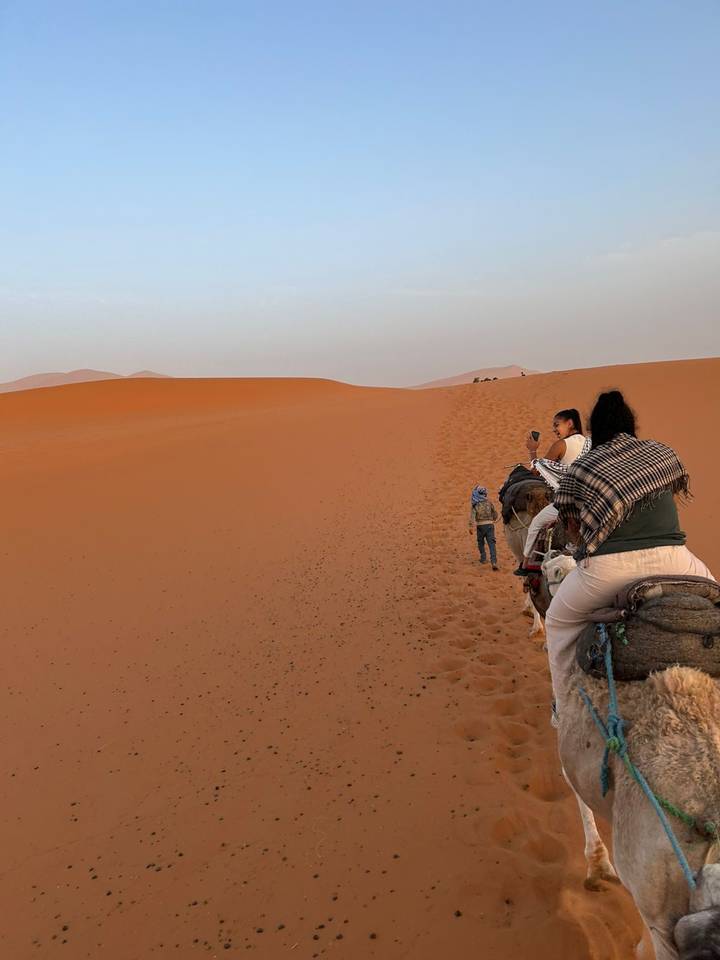 Travellers riding camels in single file across vast orange dunes at sunset in Merzouga.