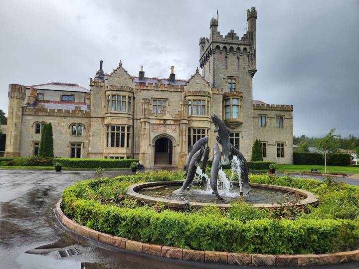Historic castle hotel with dolphin fountain surrounded by manicured gardens on a rainy day.