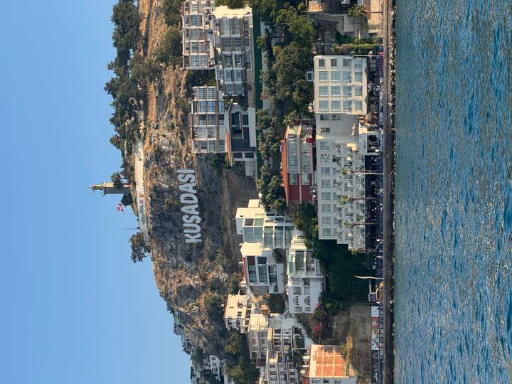 Seaside hillside with large white 'KUSADASI' sign above a coastal town and harbor under clear blue sky.