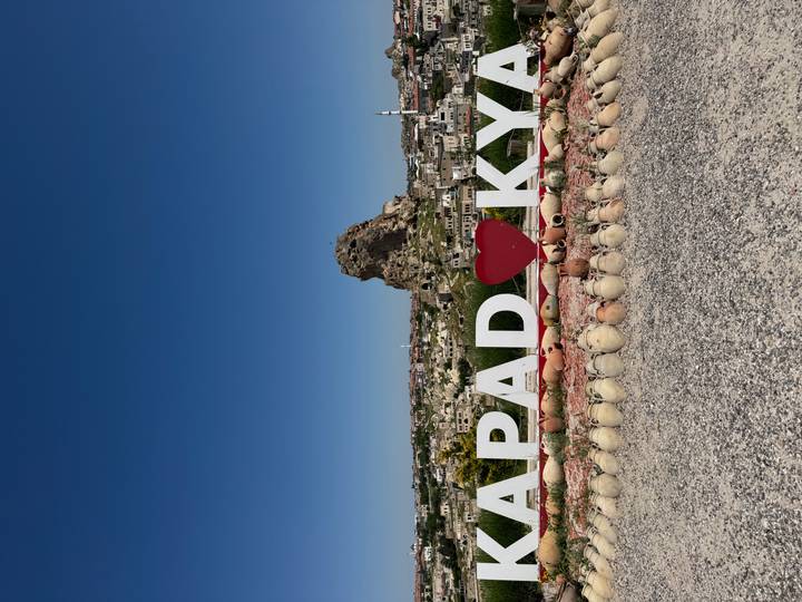 Panoramic view of a rocky town with large Cappadocia sign and heart in front of a distinctive outcrop under bright blue sky.