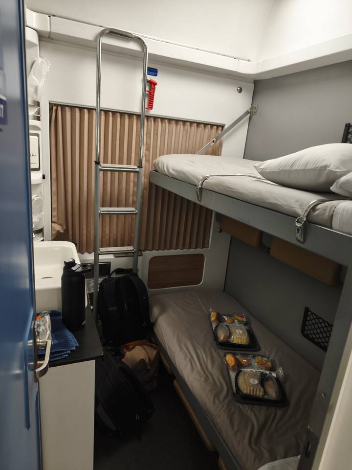 Compact sleeper cabin with bunk beds, ladder, backpack and boxed meal on lower berth.