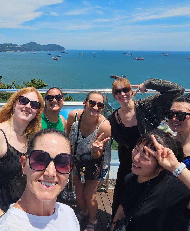 Smiling group selfie of women posing with peace signs on a seaside observation deck.