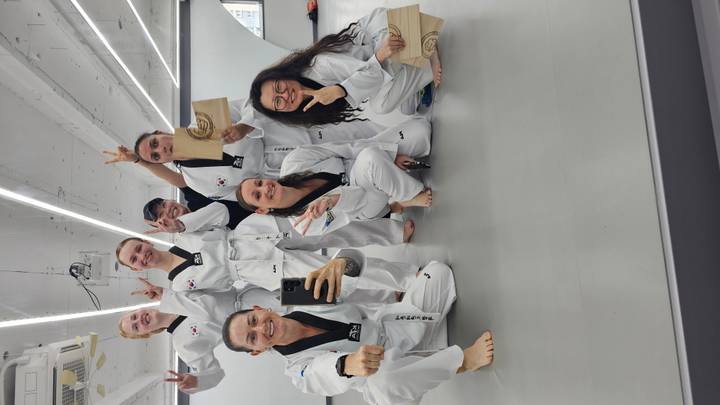 Seven participants in taekwondo uniforms pose indoors, holding broken boards and making victory signs.