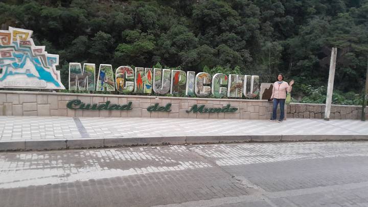 Traveler stands beside the colorful Machu Picchu welcome sign along a quiet tiled street.