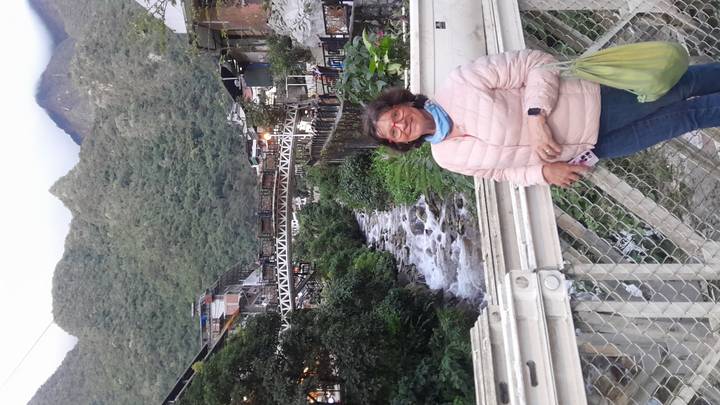 Traveler smiles on a bridge above a flowing mountain stream with town buildings and lush hills behind.