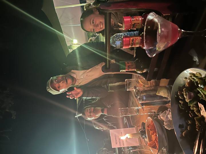 Friends enjoying drinks and food around a well-lit outdoor restaurant table at night.