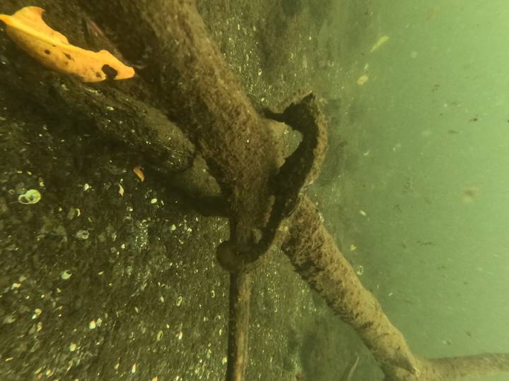 Underwater view of a rusted anchor resting on the seabed amid dark volcanic sand.
