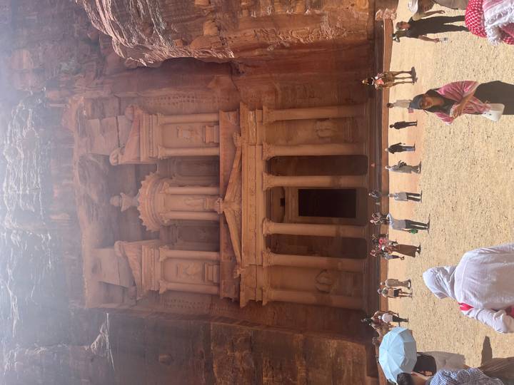 The iconic rock-carved Treasury at Petra towers above a dusty courtyard filled with tourists and camels.