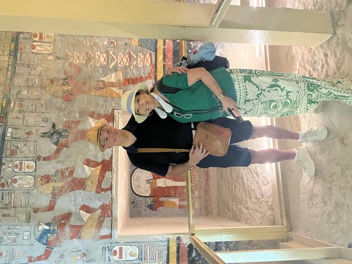 Couple poses in front of brightly painted walls showing Egyptian gods inside a sandy-floored tomb chamber.