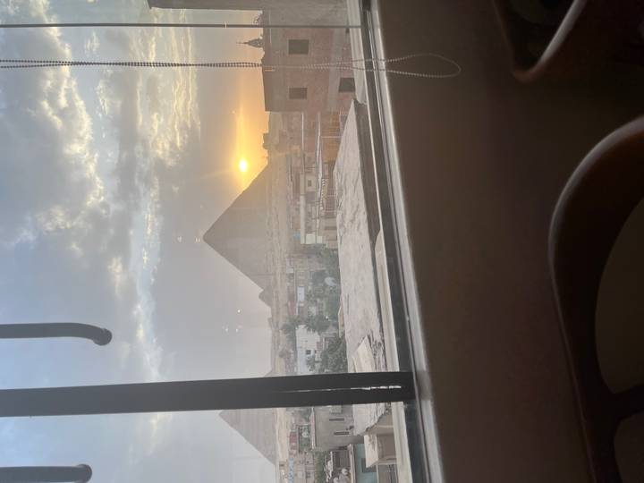 Sun sets behind the Giza Pyramid viewed through a bus window over city rooftops.
