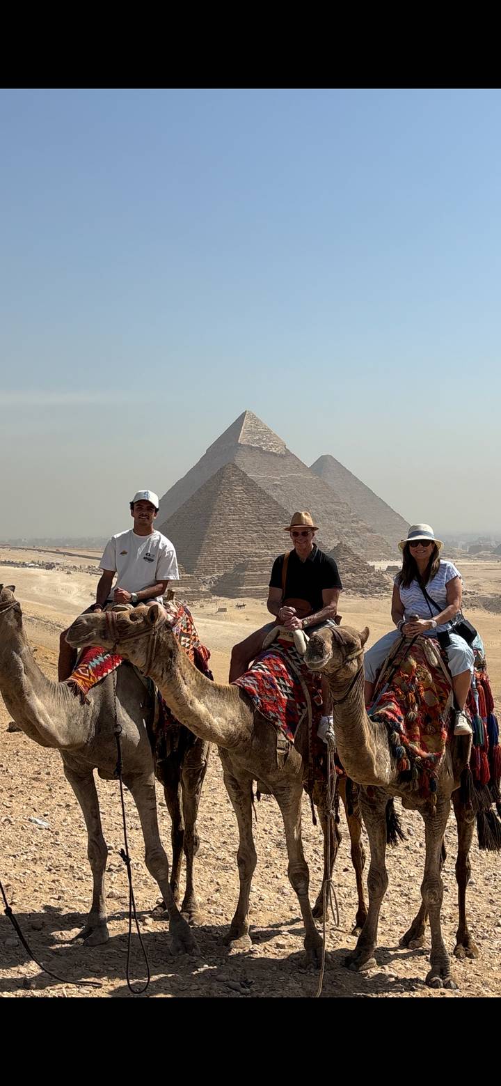 Three travelers on camels smile with the pyramids towering behind them in hazy desert light.