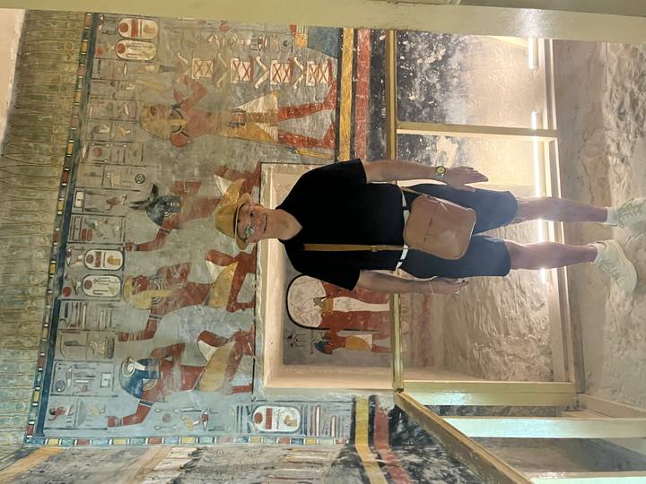 Man poses inside a richly decorated Egyptian tomb chamber with vivid wall paintings.