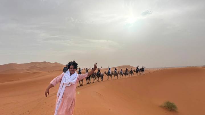 Guide flashes a peace sign as a camel caravan crosses orange Sahara dunes.