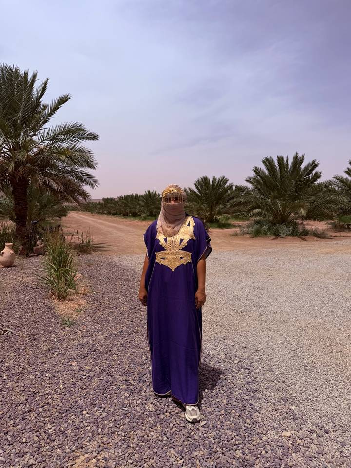 Woman in a traditional purple dress and face veil stands among desert palms.