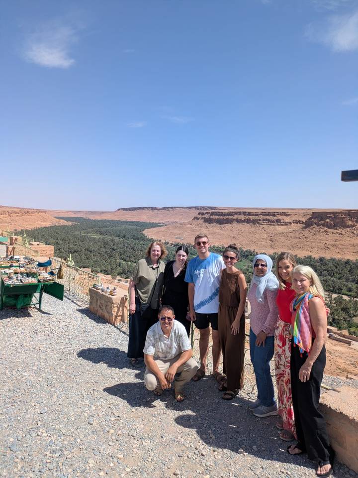 Travel group poses at a viewpoint overlooking a lush oasis valley and desert cliffs.
