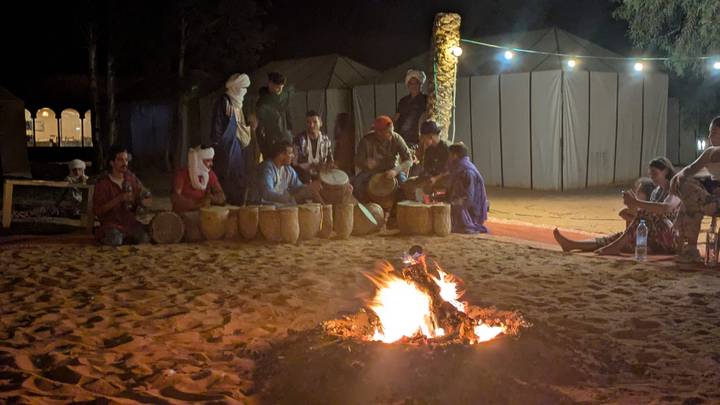 Nighttime campfire with musicians drumming and tourists relaxing in a desert camp.