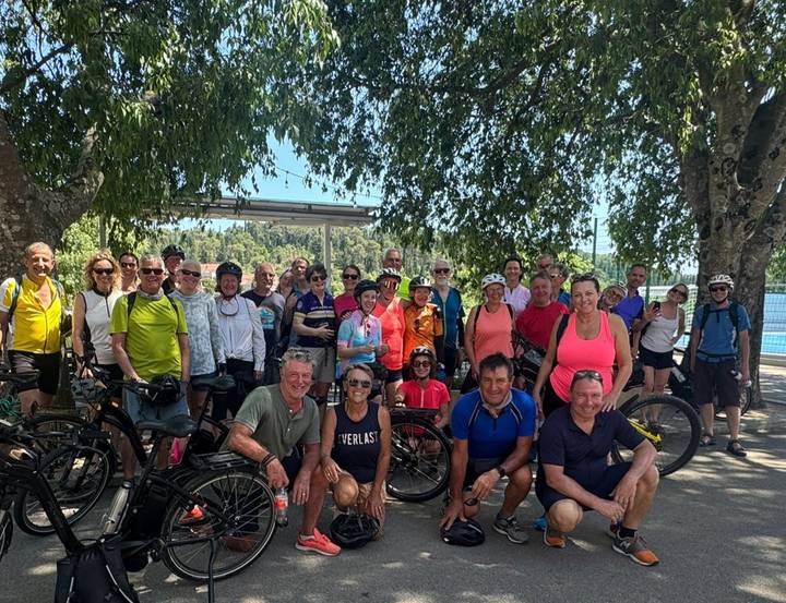 Large cycling tour group poses together with their bikes under leafy trees.