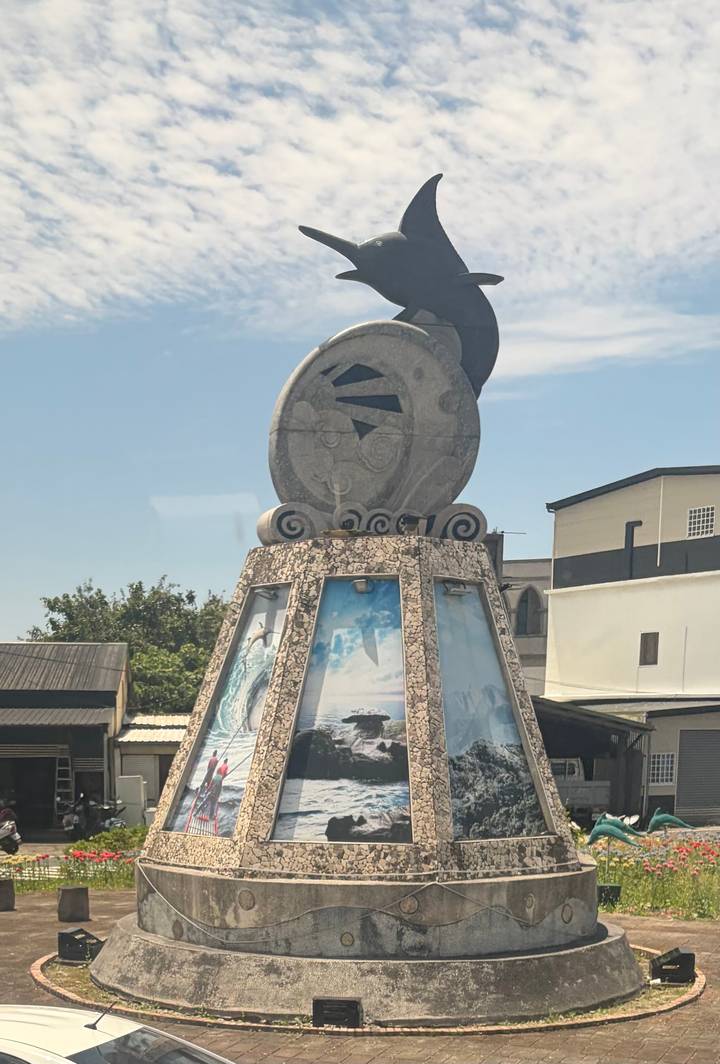A sculptural monument topped with a round stone artwork against a blue sky; photos are displayed on its tiled base.