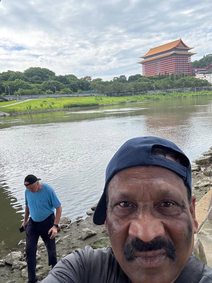 Partial selfie showing the top of a man’s head by a riverbank while another person looks at the water behind him.