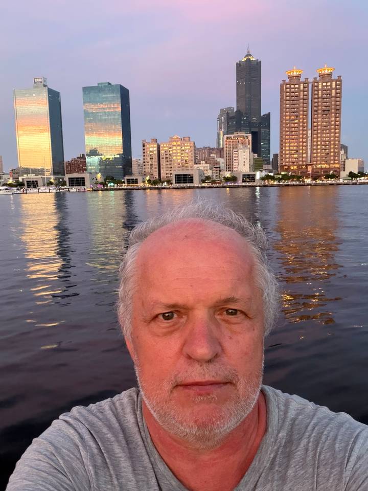 Close-up selfie of a man with a lit city waterfront skyline reflecting on calm water at dusk.