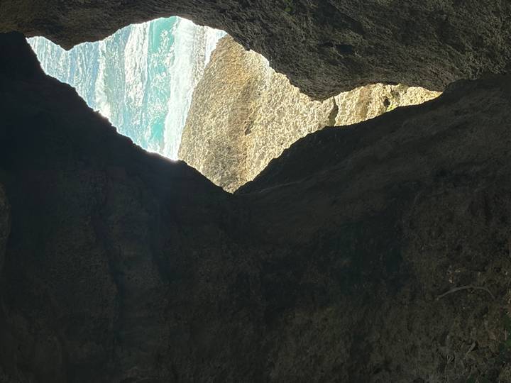 View through a dark rocky cave opening toward bright turquoise waves crashing on rocks.